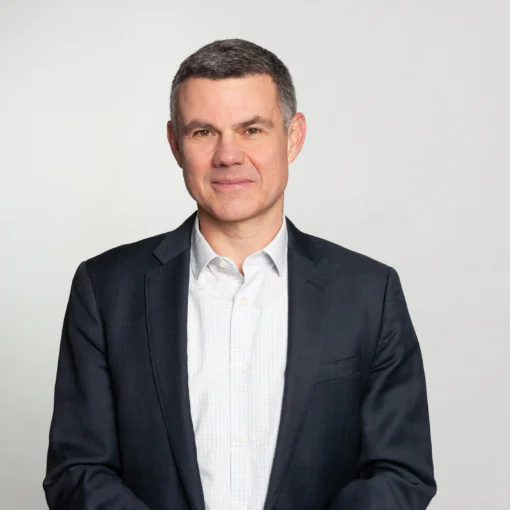 photo of a white man with gray hair in a white button up shirt and black blazer against a gray background