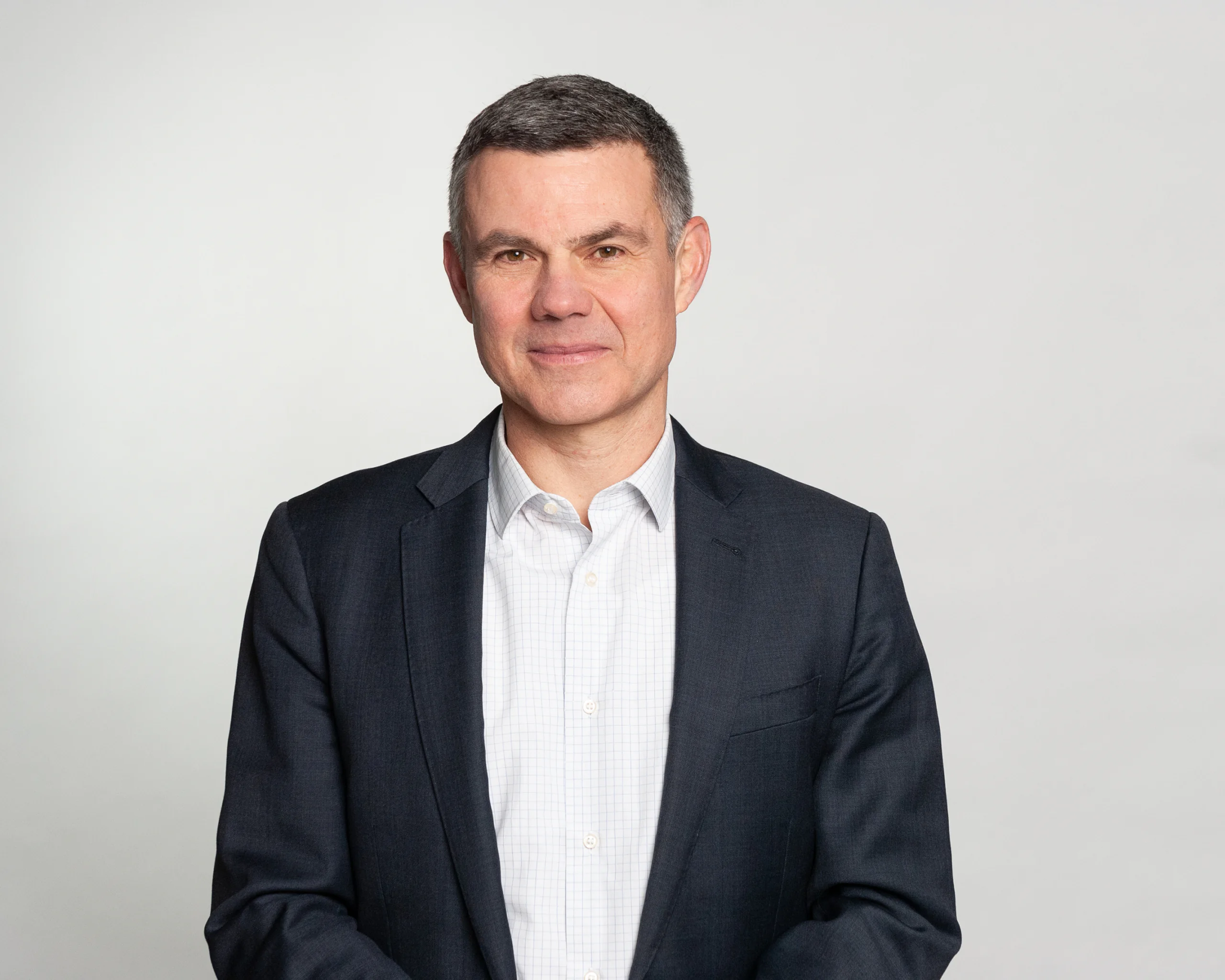 photo of a white man with gray hair in a white button up shirt and black blazer against a gray background