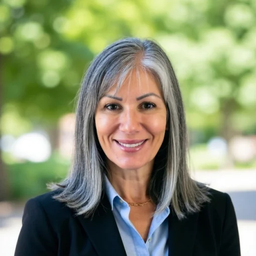white woman with gray hair wearing a blue button up shirt and black blazer against a natural green background.